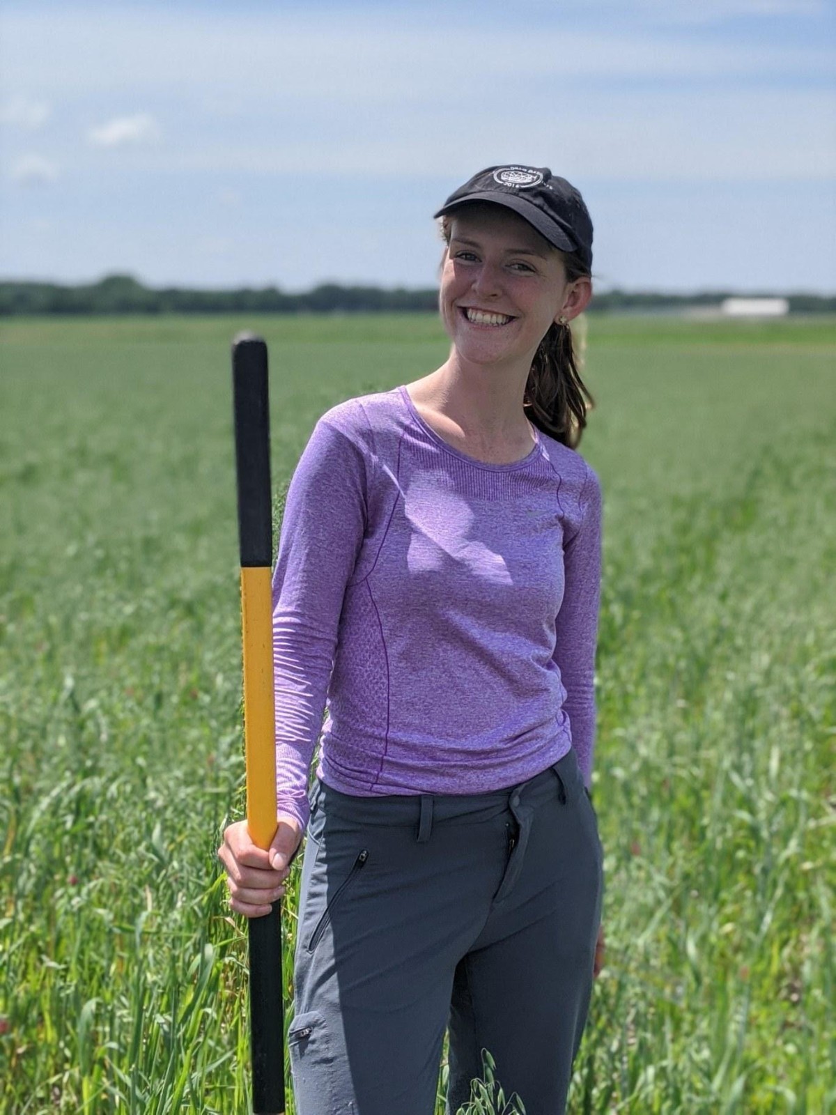 Rebecca smiling while holding a shovel and standing in a field of grassy cover crops