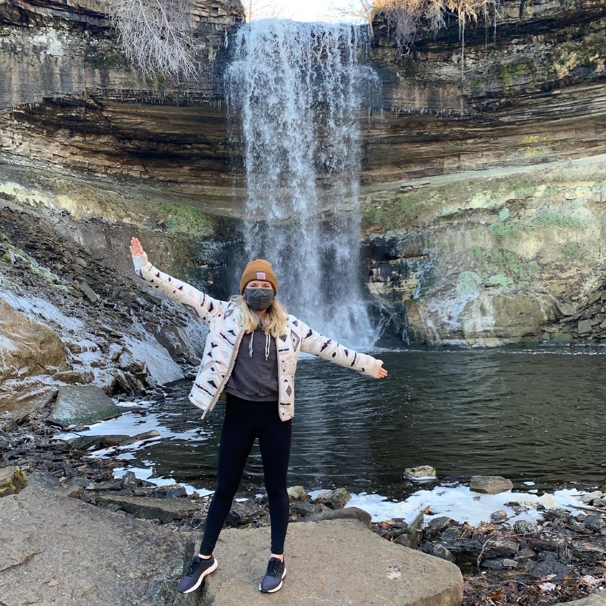 Sarah posing with her arms spread out wide in front of a waterfall and some stratified brown and green rocks. She is wearing black pants and a white jacket with a yellow winter hat and gray face mask