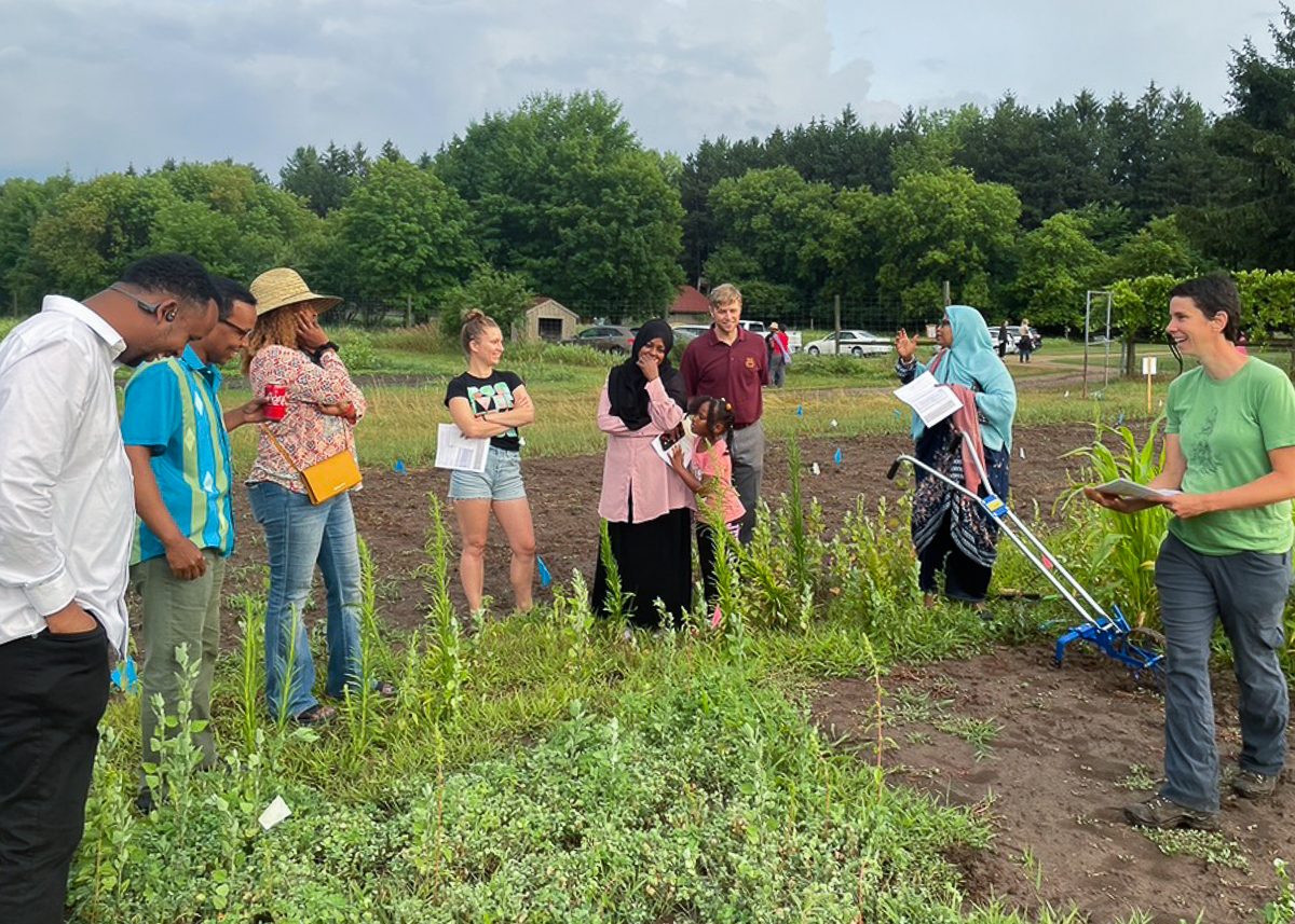 A group of students stands and listens to a researcher speaking to them in a field of greens